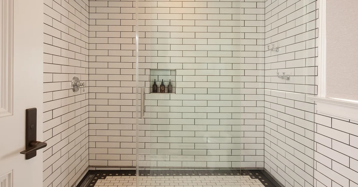 A shower with white subway tile, a wall niche, wall controls and a glass enclosure.
