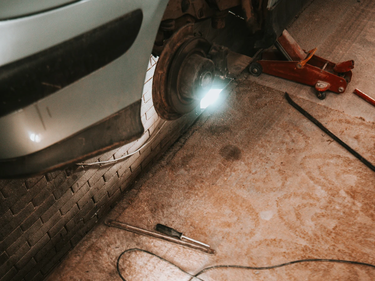 Car in a workshop with a wheel removed and a portable work light on the floor.