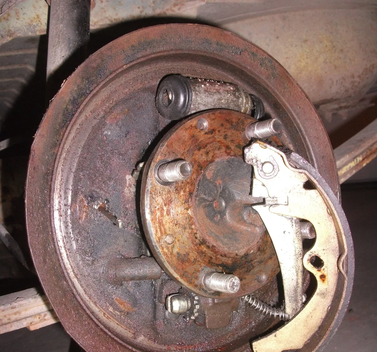 A rusty rear drum brake with the drum taken off, showing the backing plate, springs and one brake shoe