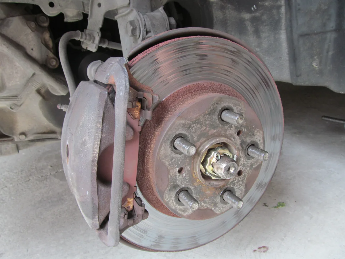 An old car front brake with the wheel off, showing a rusty caliper over a grooved brake rotor